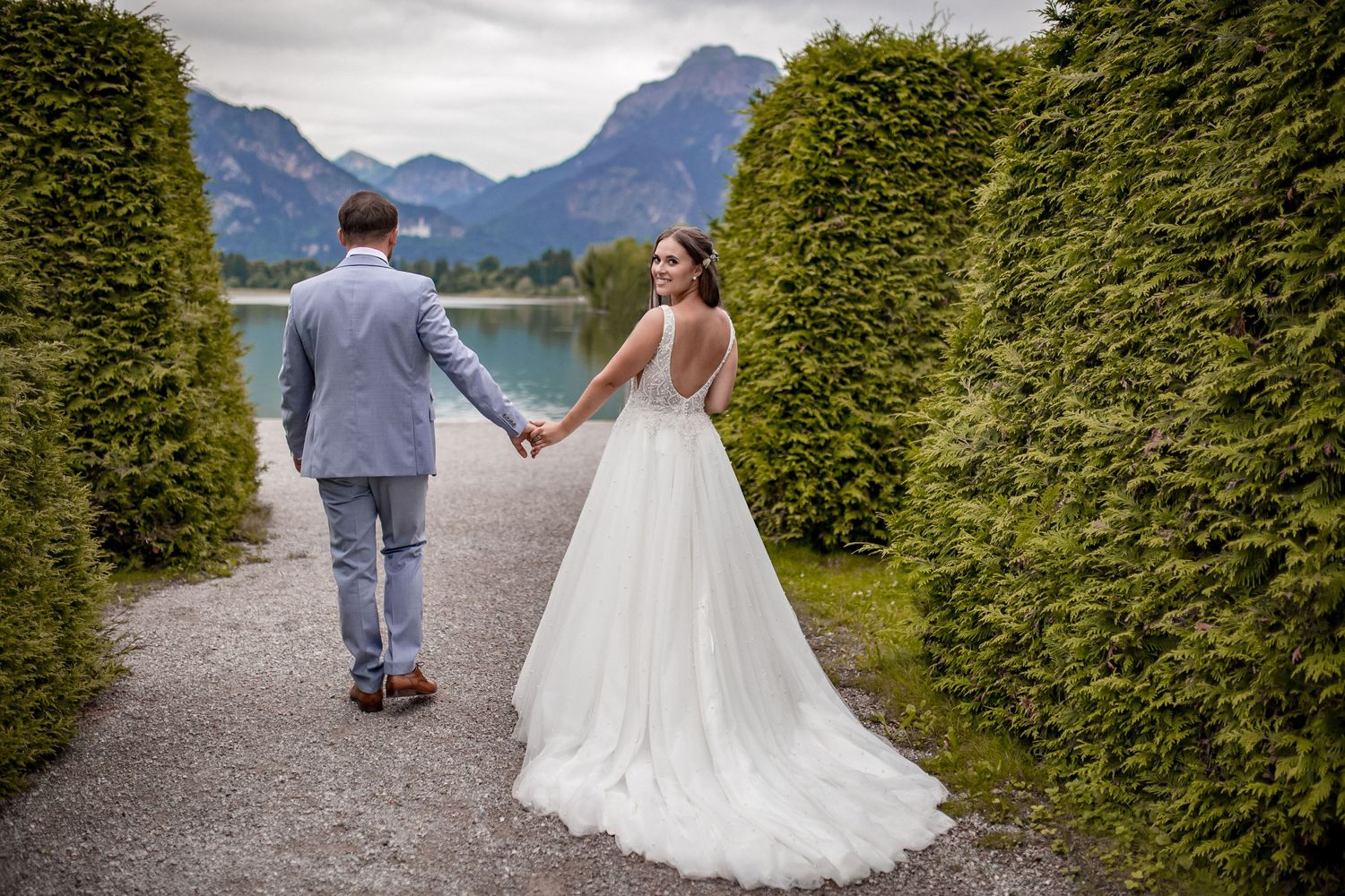 Couple walking hand in hand on path between tall hedges with mountains in background.