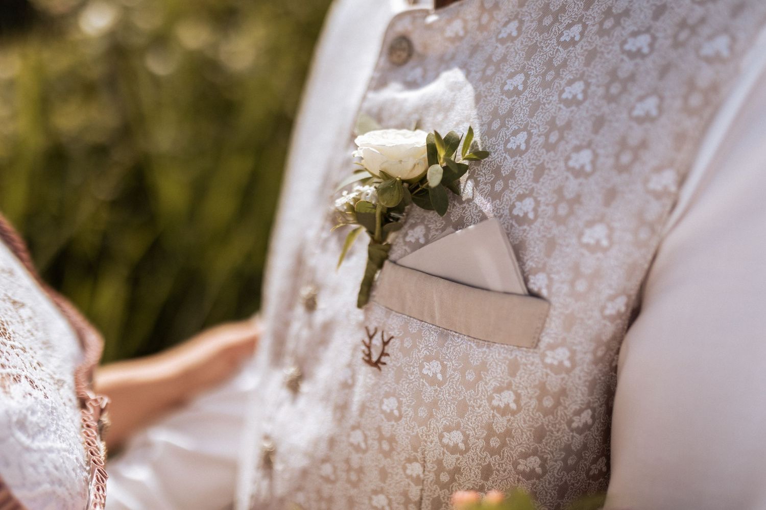 A white boutonniere pinned to a light colored vest with water droplets on the fabric.