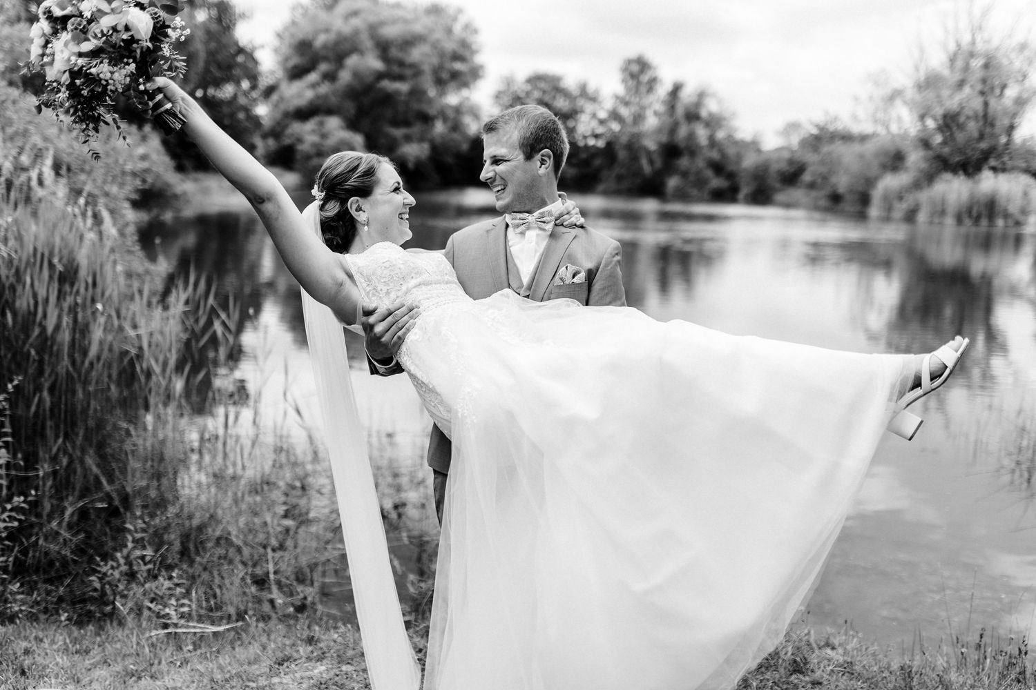 Black and white photo of newlyweds in romantic pose by a lake with bride being lifted.