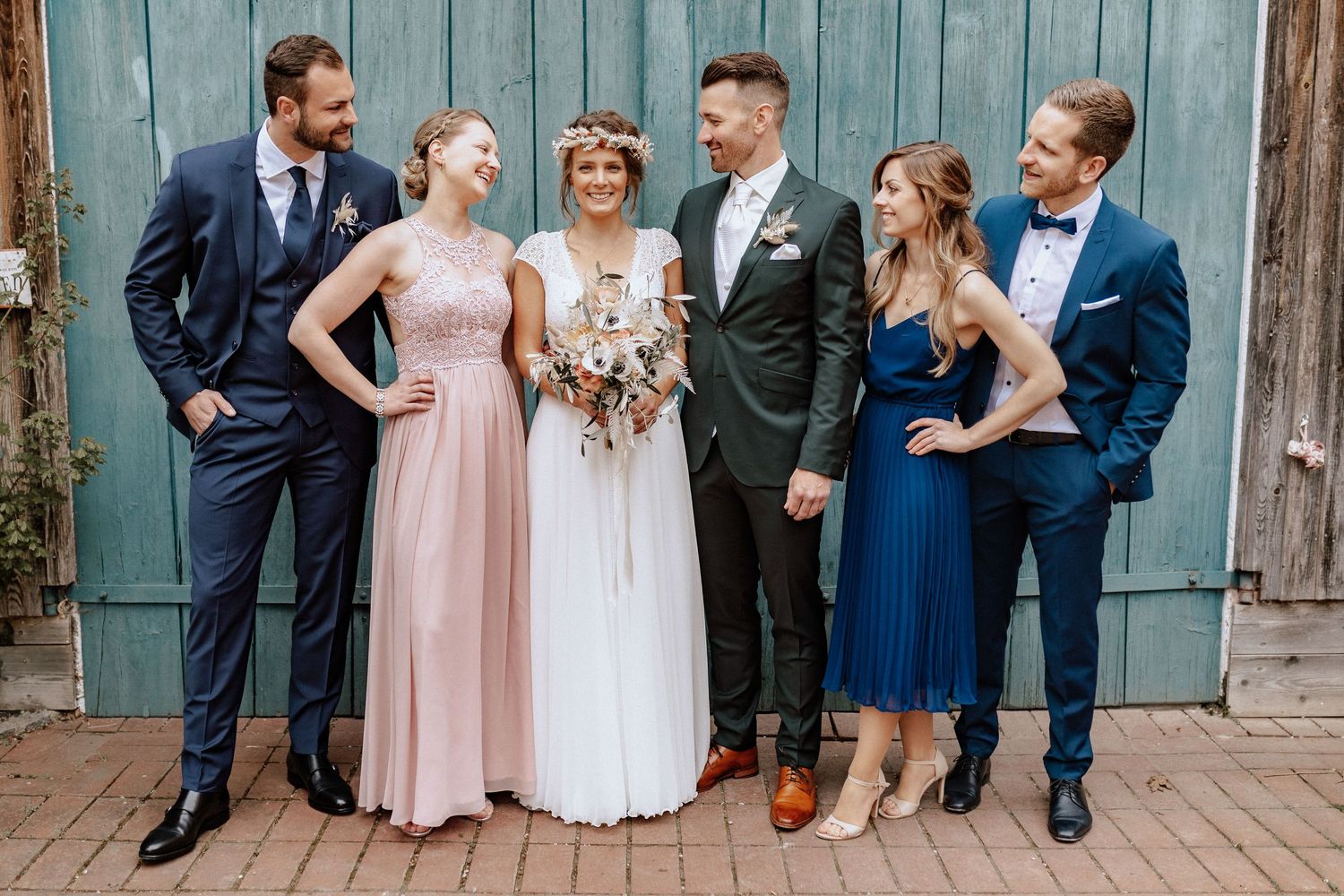 Wedding party of six poses in formal attire against weathered barn door.