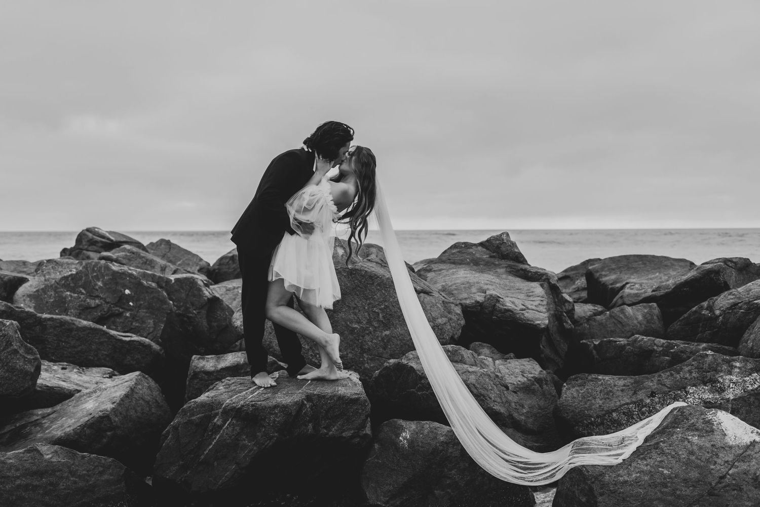 A dramatic black and white wedding photo with a flowing veil on rocky coastal cliffs.