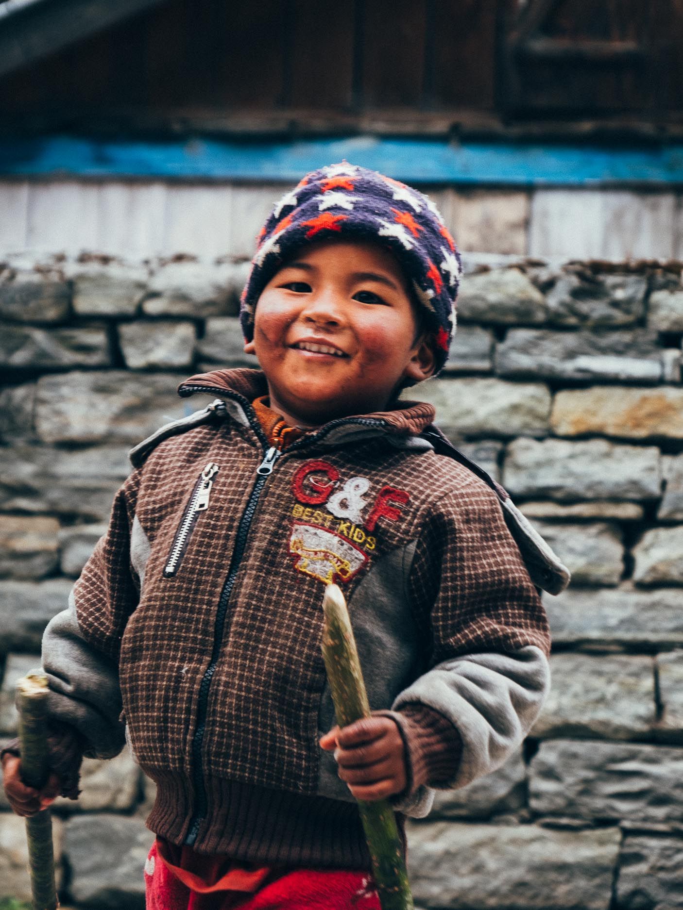 A child in winter clothing smiles brightly while standing against a stone wall in Nepal.