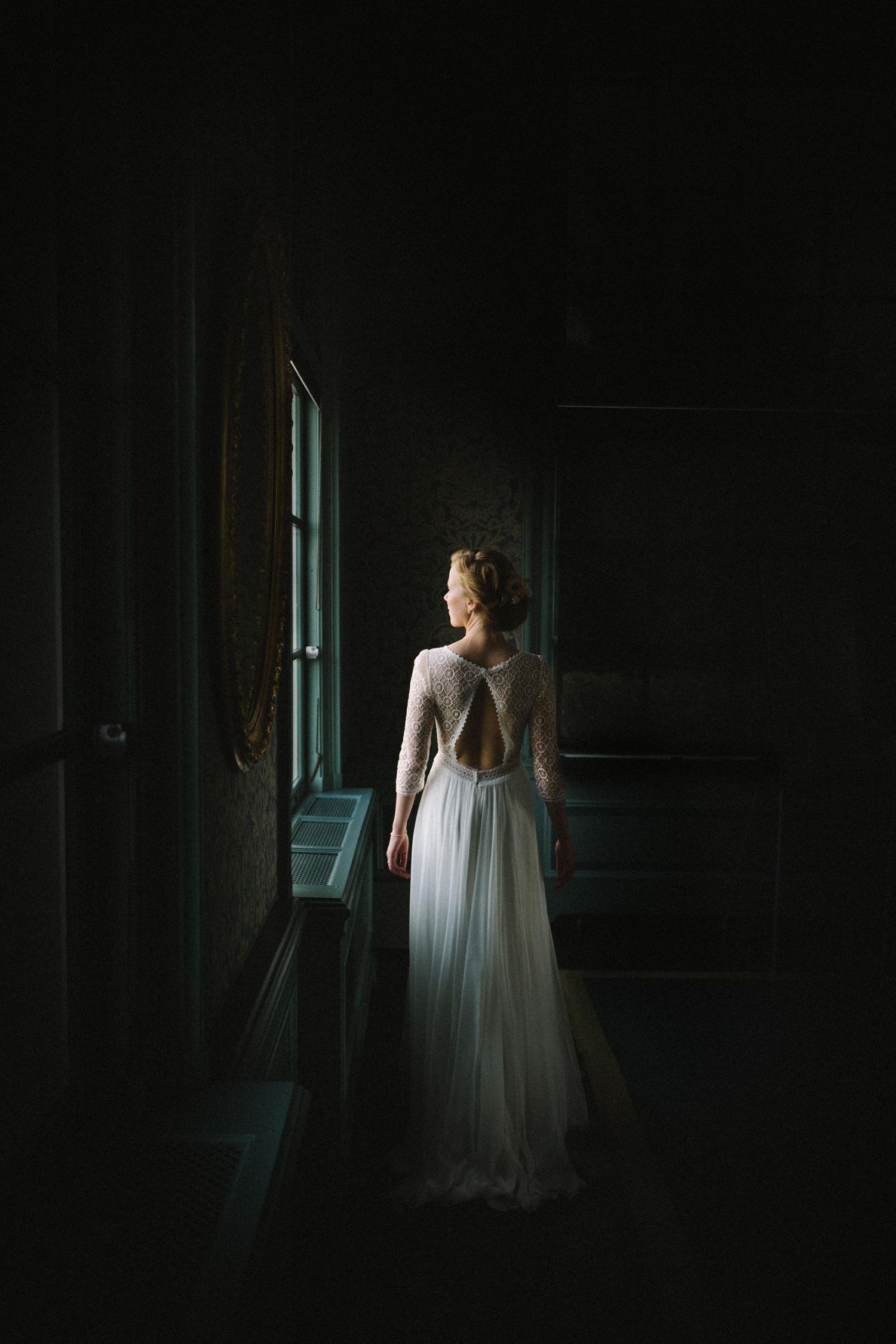 Dramatic bridal portrait in dark room with natural window light highlighting wedding dress.