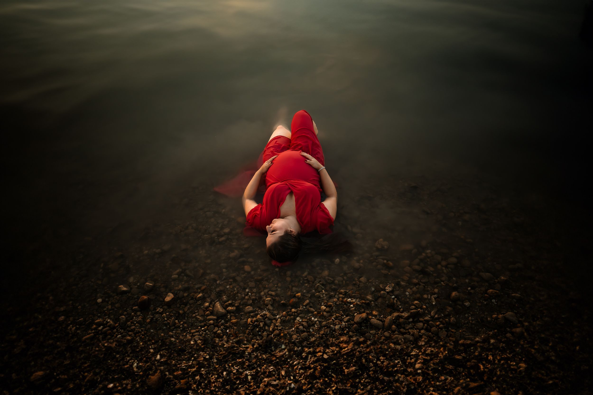 A person in red lies on dark pebbles at the water's edge creating a moody scene.