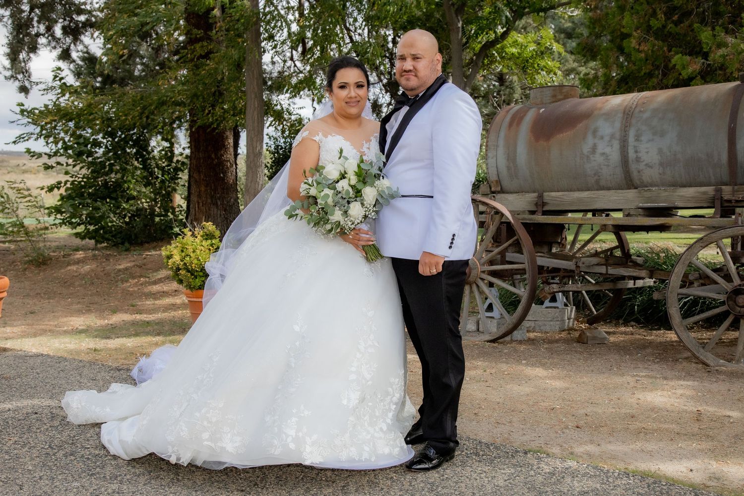 Bride and groom pose together in elegant wedding attire near rustic wagon and trees outdoors.