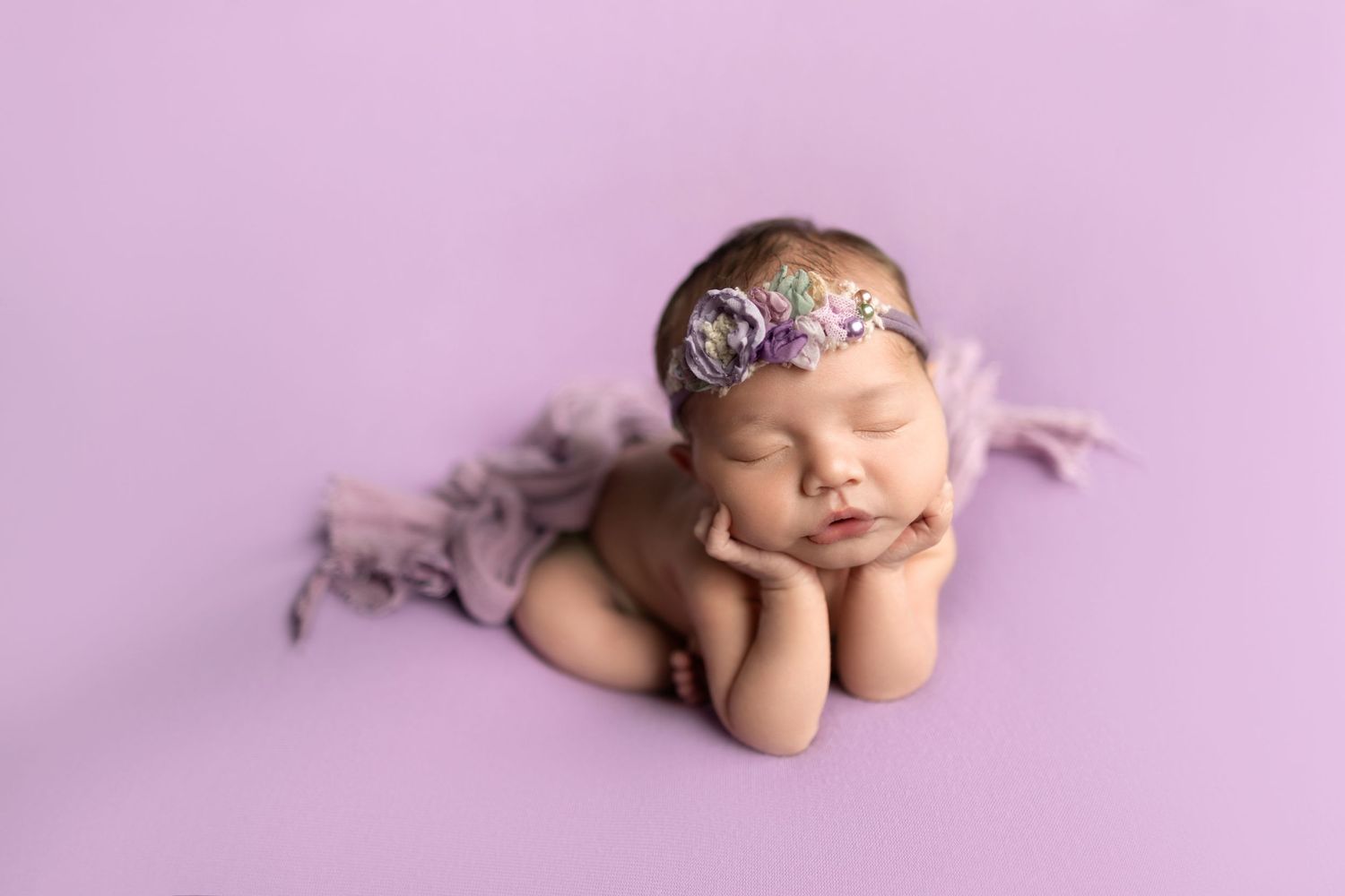 Newborn baby sleeping peacefully with floral headpiece against purple backdrop.