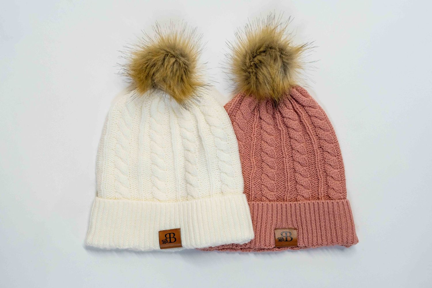 Two knit winter beanies in white and pink with faux fur pom poms.