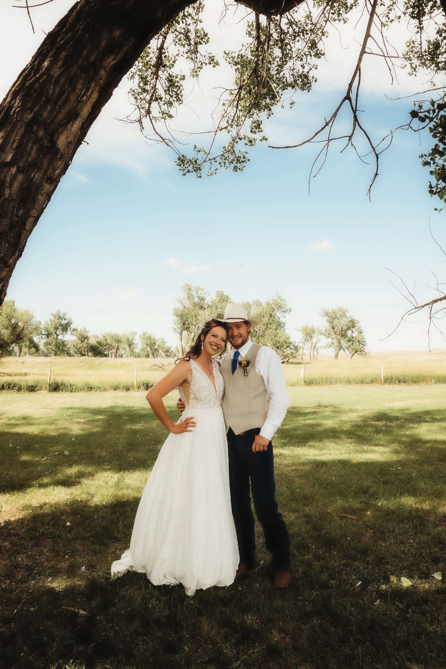 A rustic outdoor wedding photo featuring newlyweds standing under a large tree.