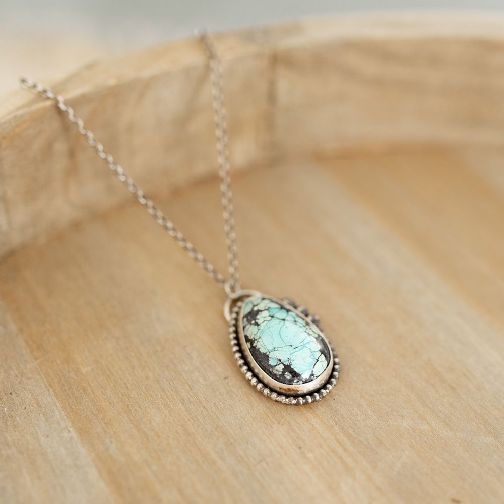 Delicate turquoise pendant necklace with silver detailing on beige textured surface.