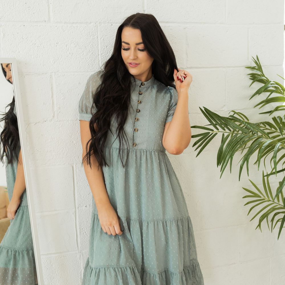 A sage green tiered midi dress with short puff sleeves and button details photographed against a white wall.