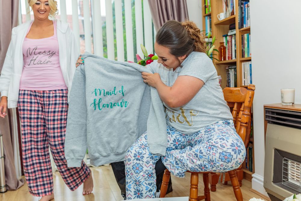 bridesmaid holds up her wedding git from the bride a sweater with maid of honour printed on it