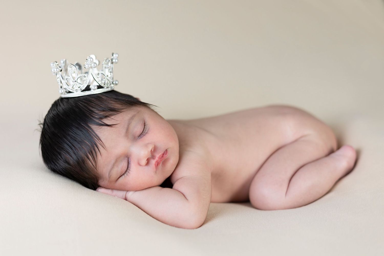 Newborn baby sleeping wearing a tiny silver crown in gentle lighting.