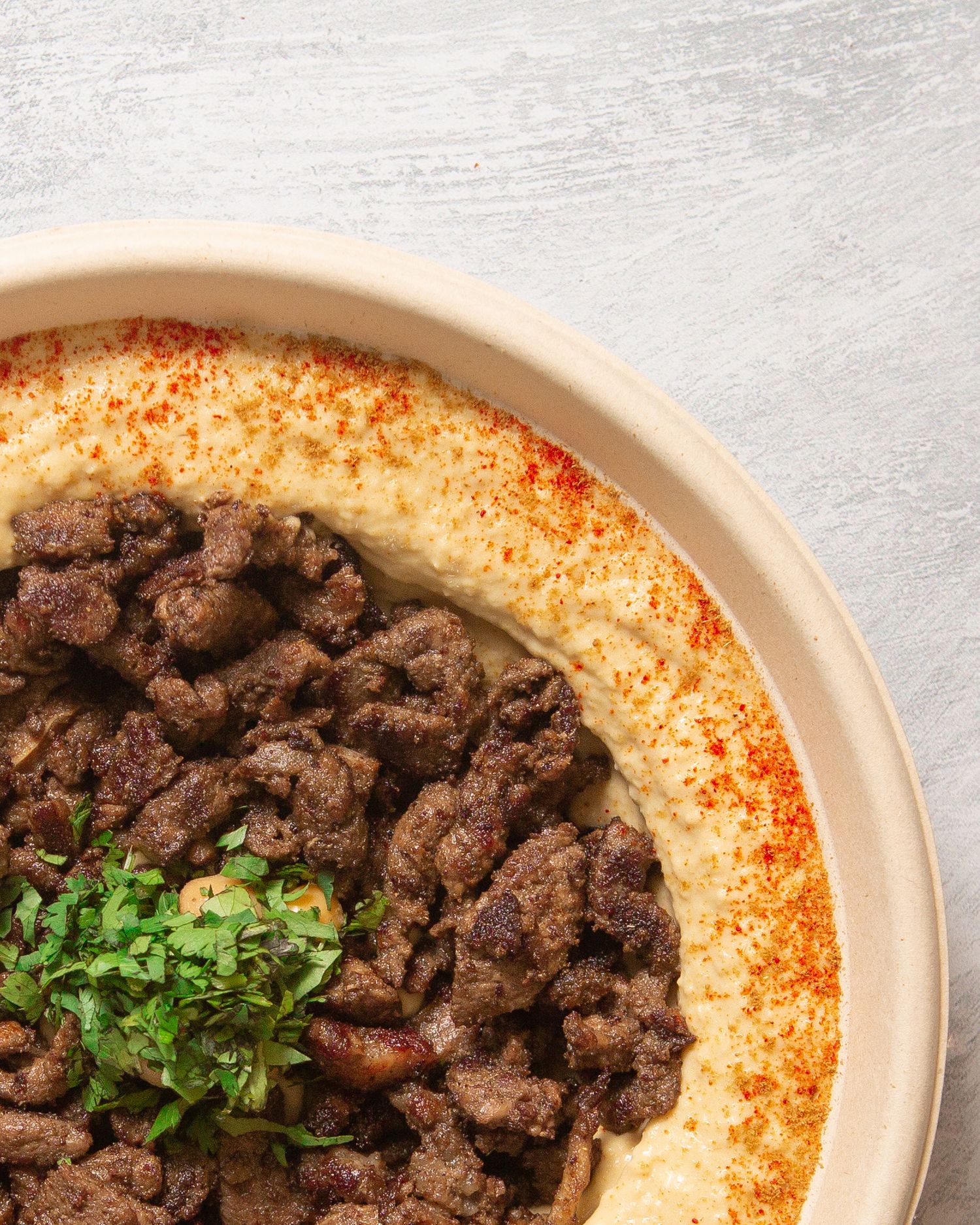 A bowl of hummus sprinkled with paprika and garnished with fresh herbs and spiced meat.