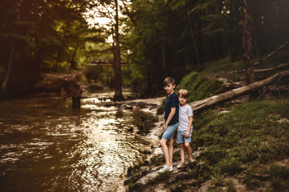 Two people stand together on rocks beside a flowing river in a forest setting.