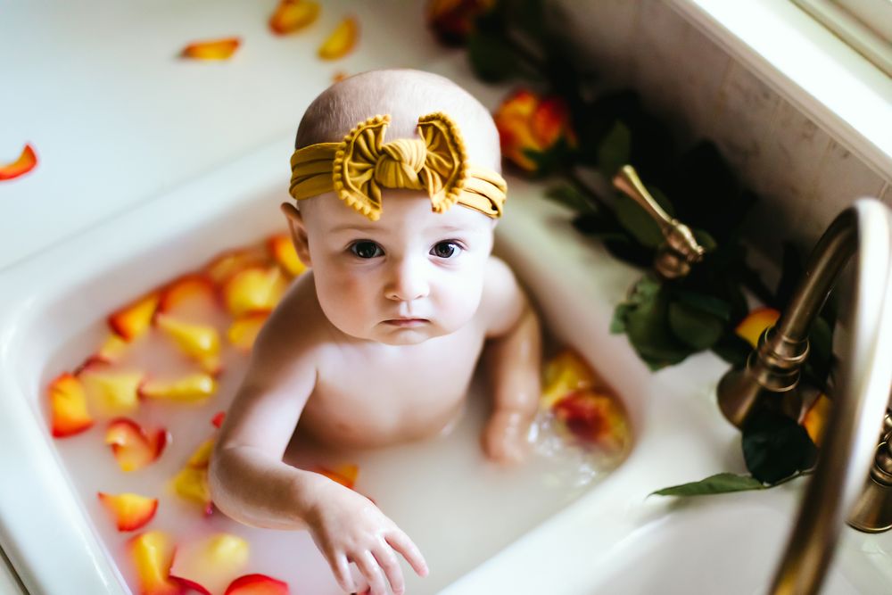 Baby wearing gold headband in bathtub surrounded by orange flower petals.