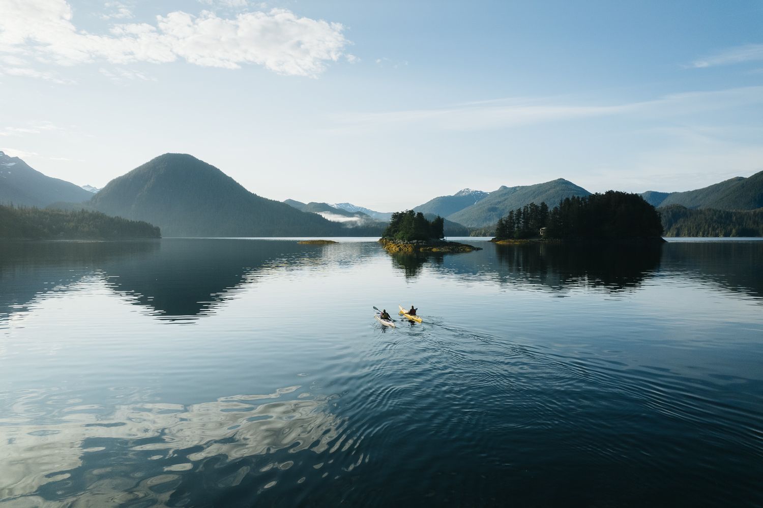 Two kayakers paddle on a serene mountain lake surrounded by lush peaks and reflective waters at dawn.