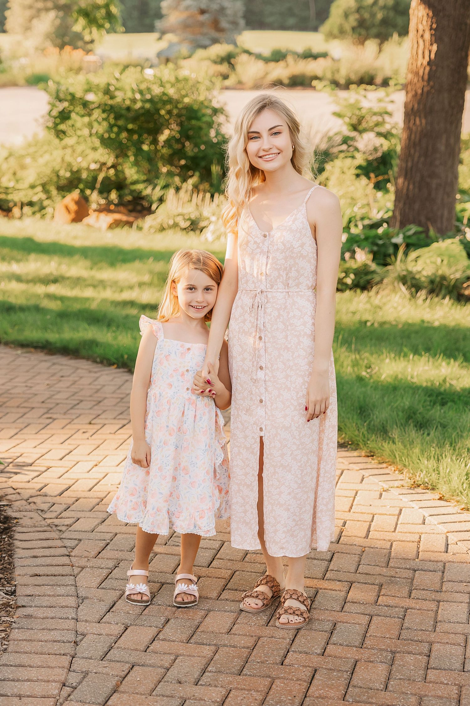 Two people in light pink dresses pose together on brick pathway in garden setting.