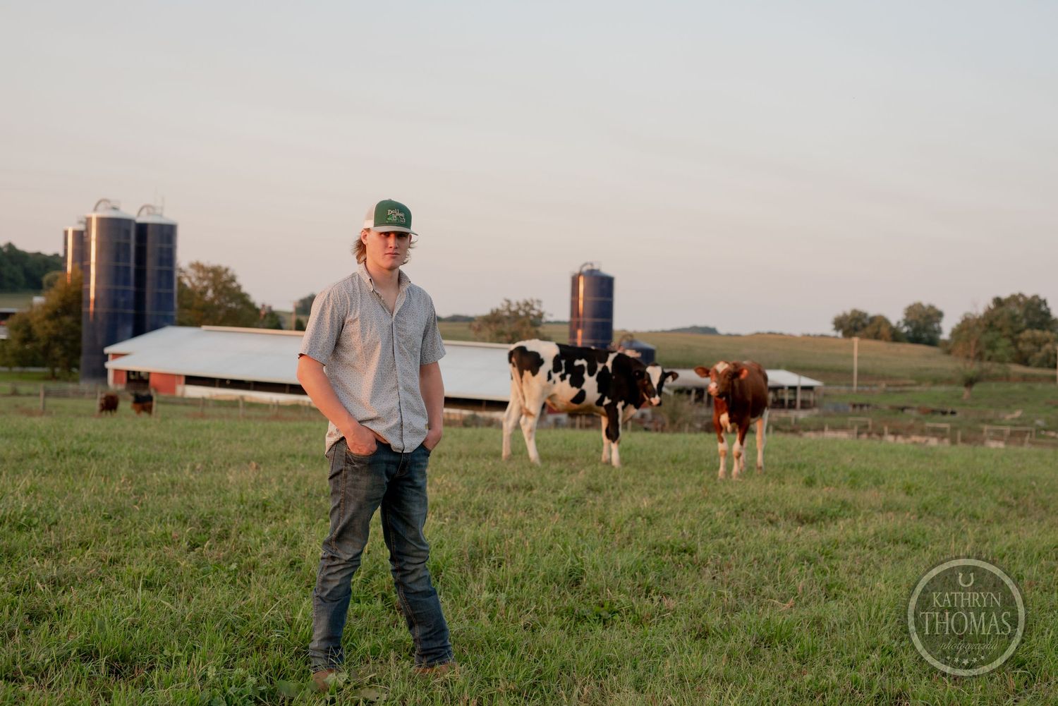 Farmer stands in pasture with dairy cows and farm buildings visible in background at dusk.