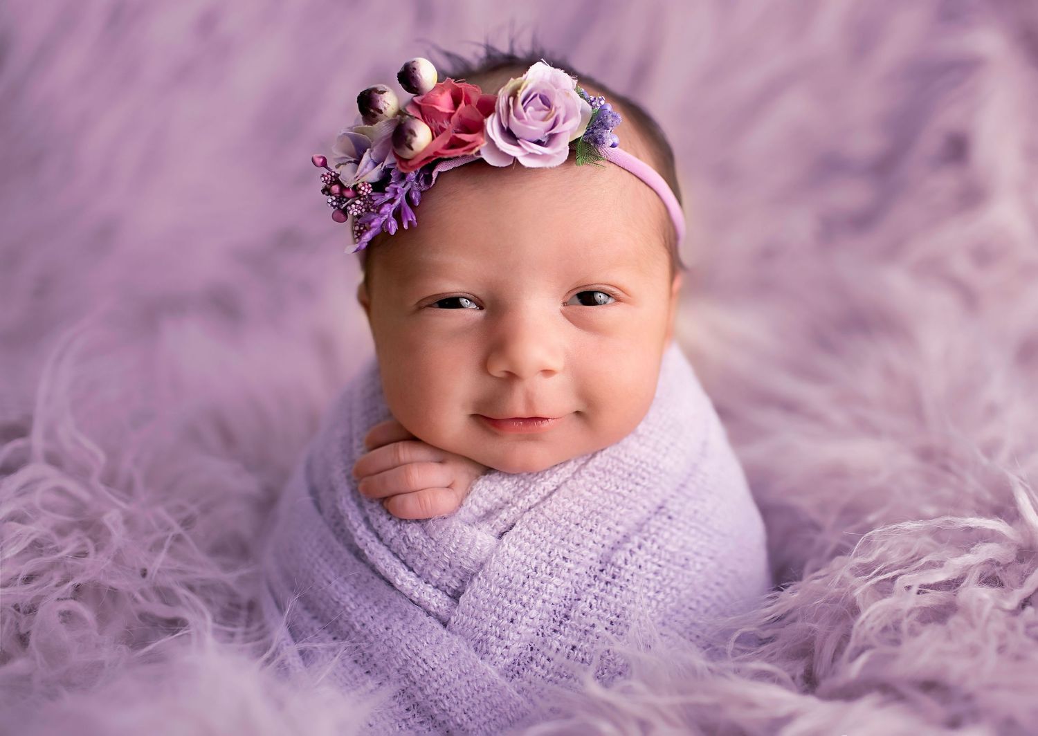 Sweet baby wrapped in purple knit blanket wearing floral headband on fluffy purple background.