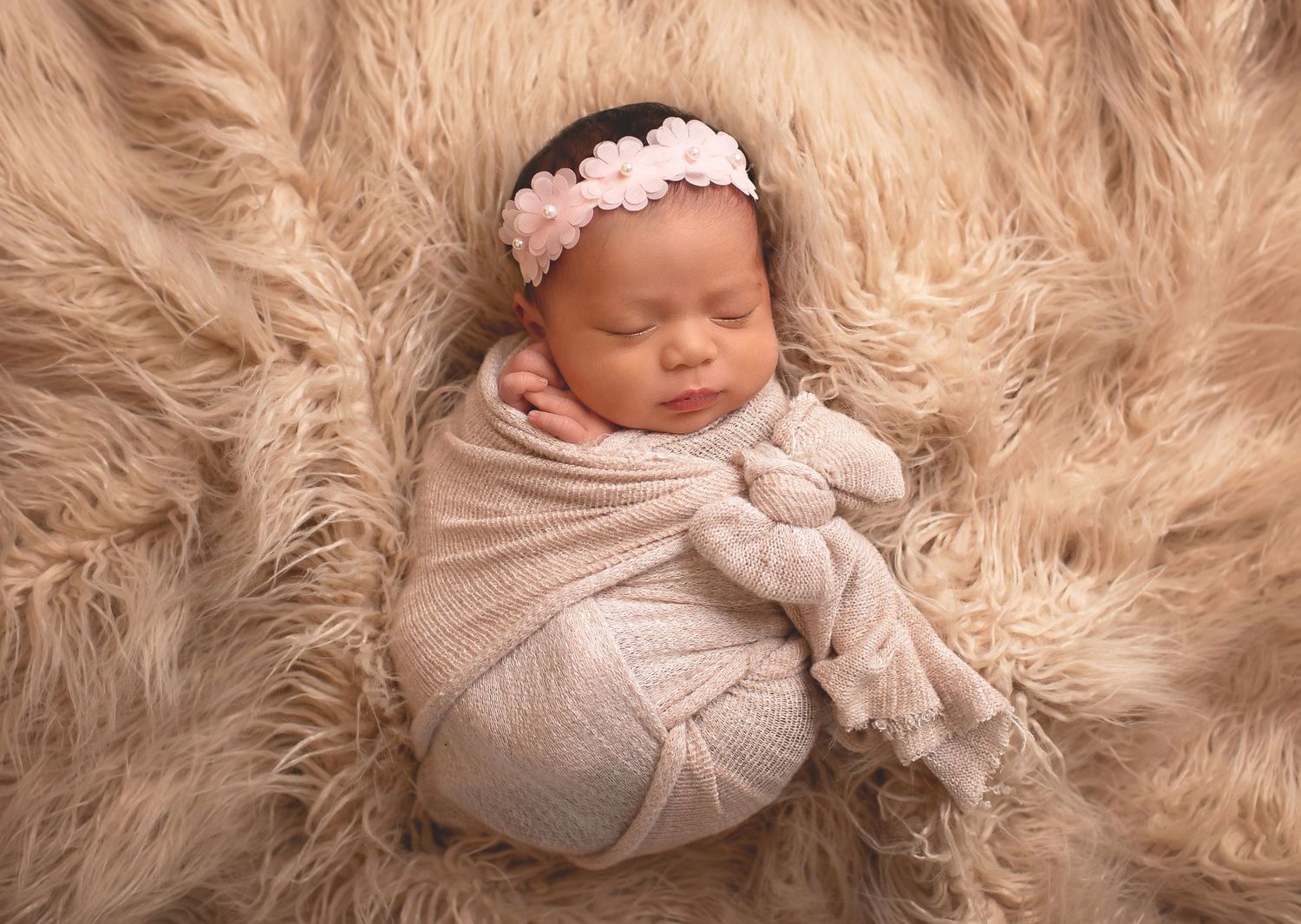 Sleeping newborn wrapped in soft beige knit swaddle with lace headband resting on fluffy taupe fur backdrop.