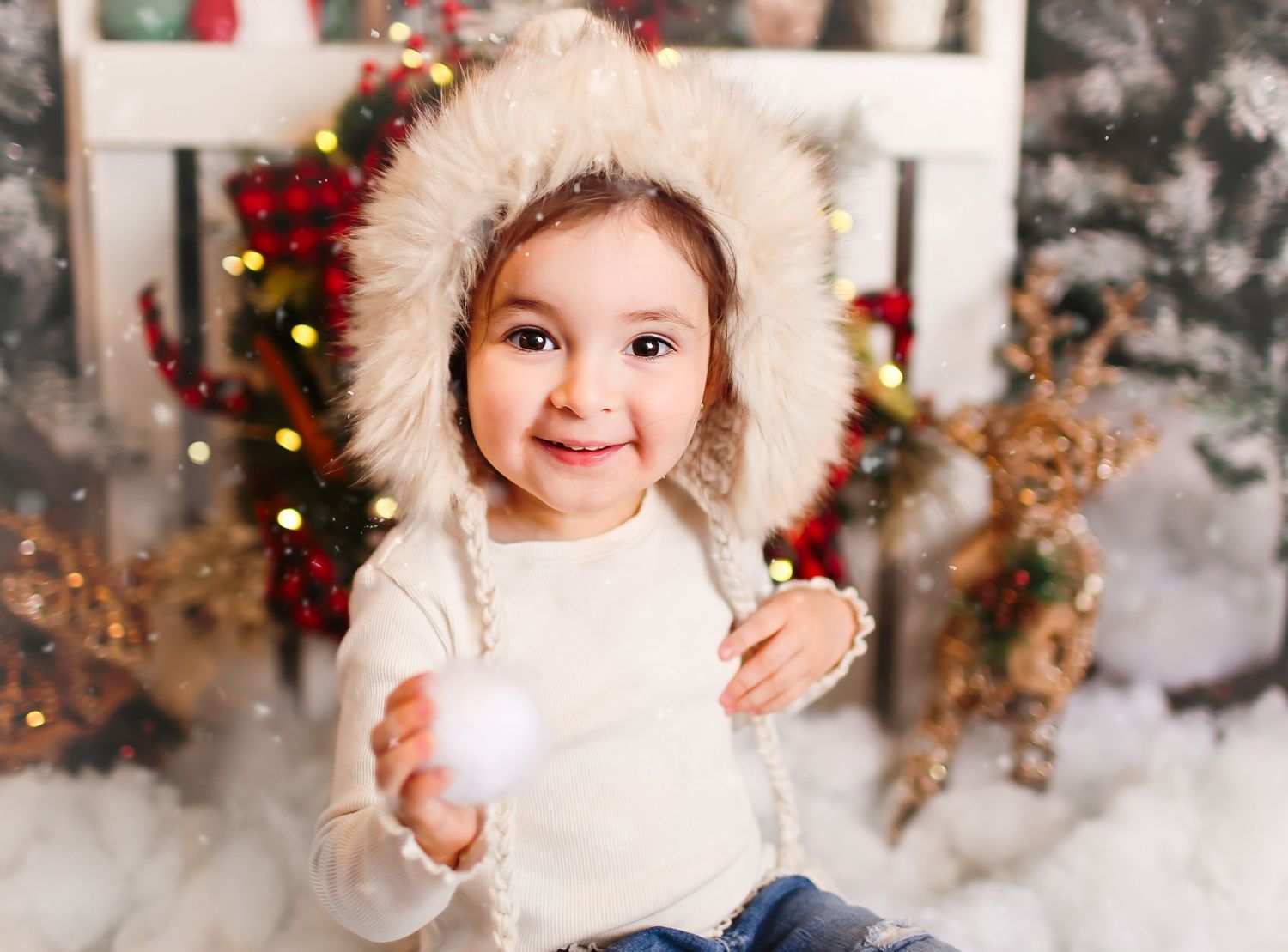 A toddler wearing a white furry hood holds a snowball in a festive holiday setting with Christmas decorations.