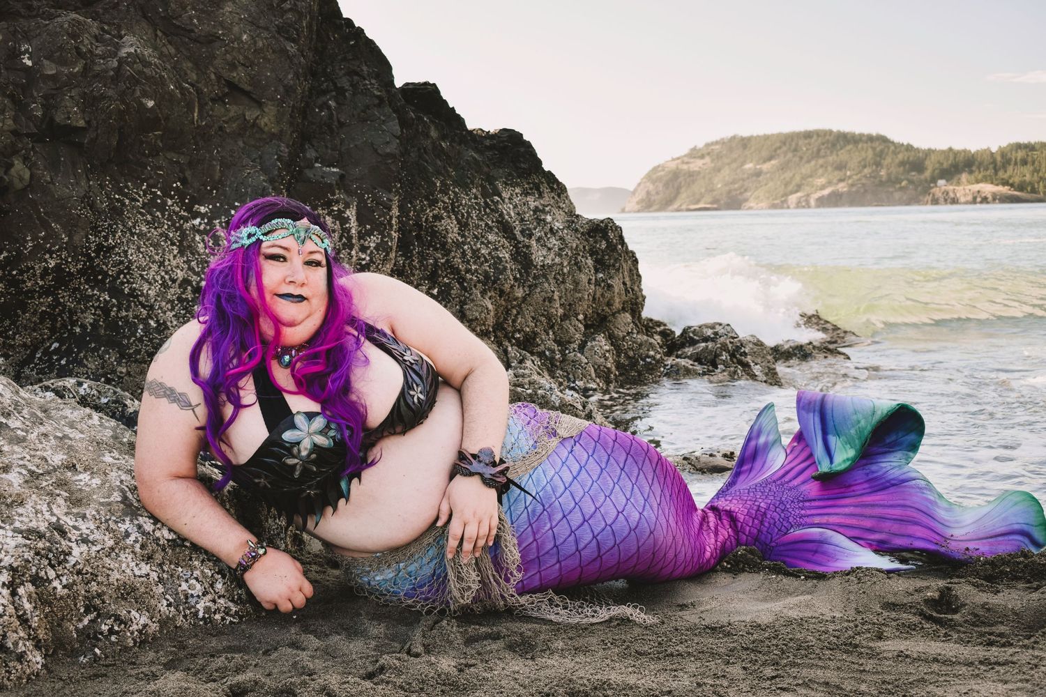 Person in purple mermaid costume poses dramatically on rocky beach at sunset.