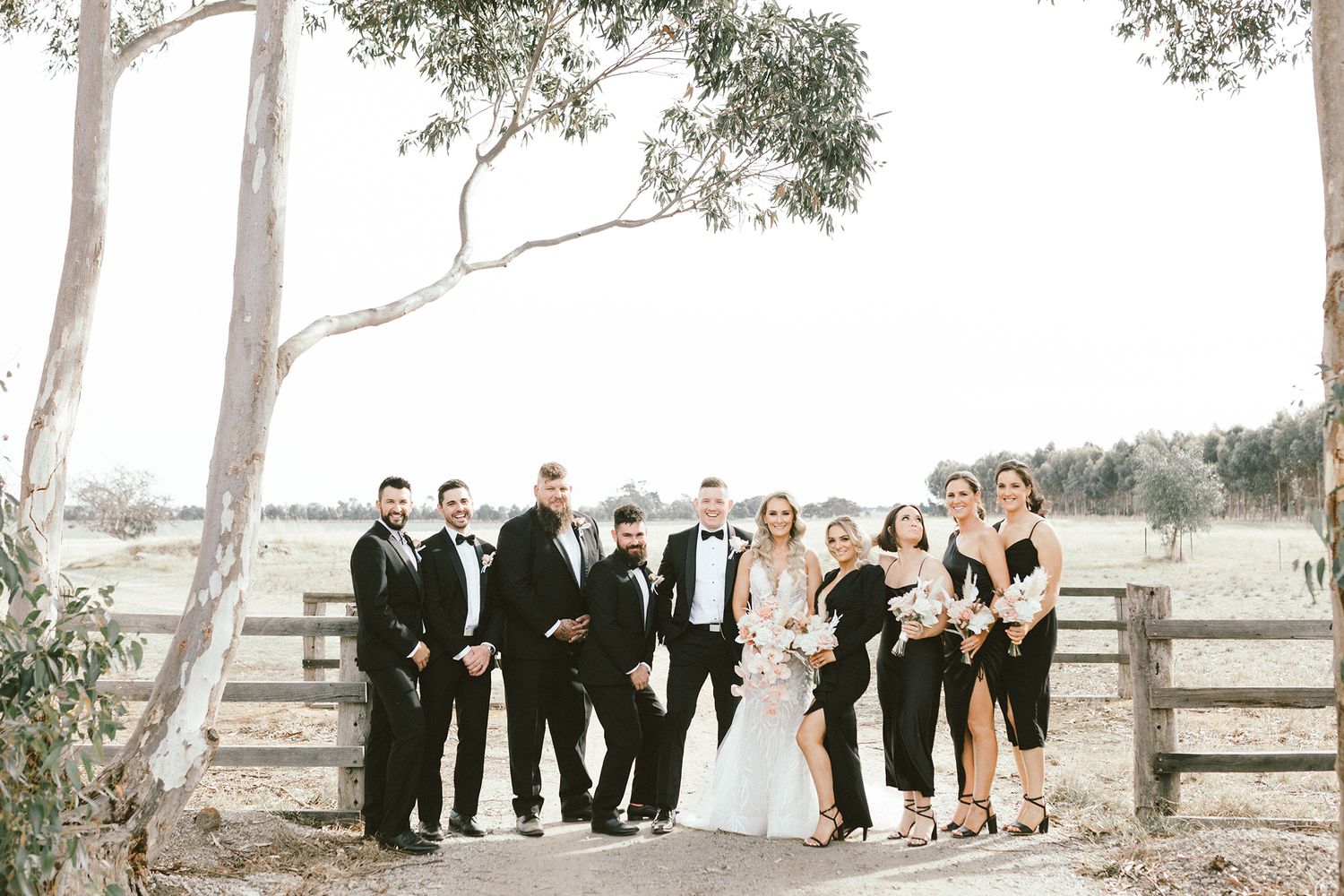 Wedding group poses by rustic wooden fence with eucalyptus trees in background.
