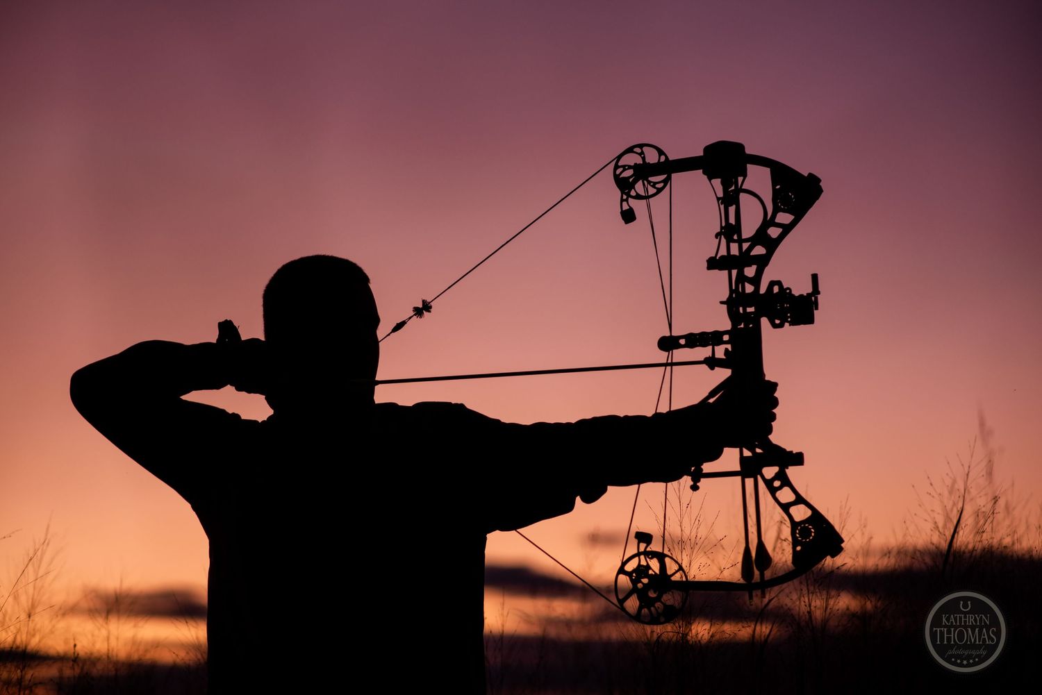 Silhouette of archer drawing compound bow against dramatic sunset sky.