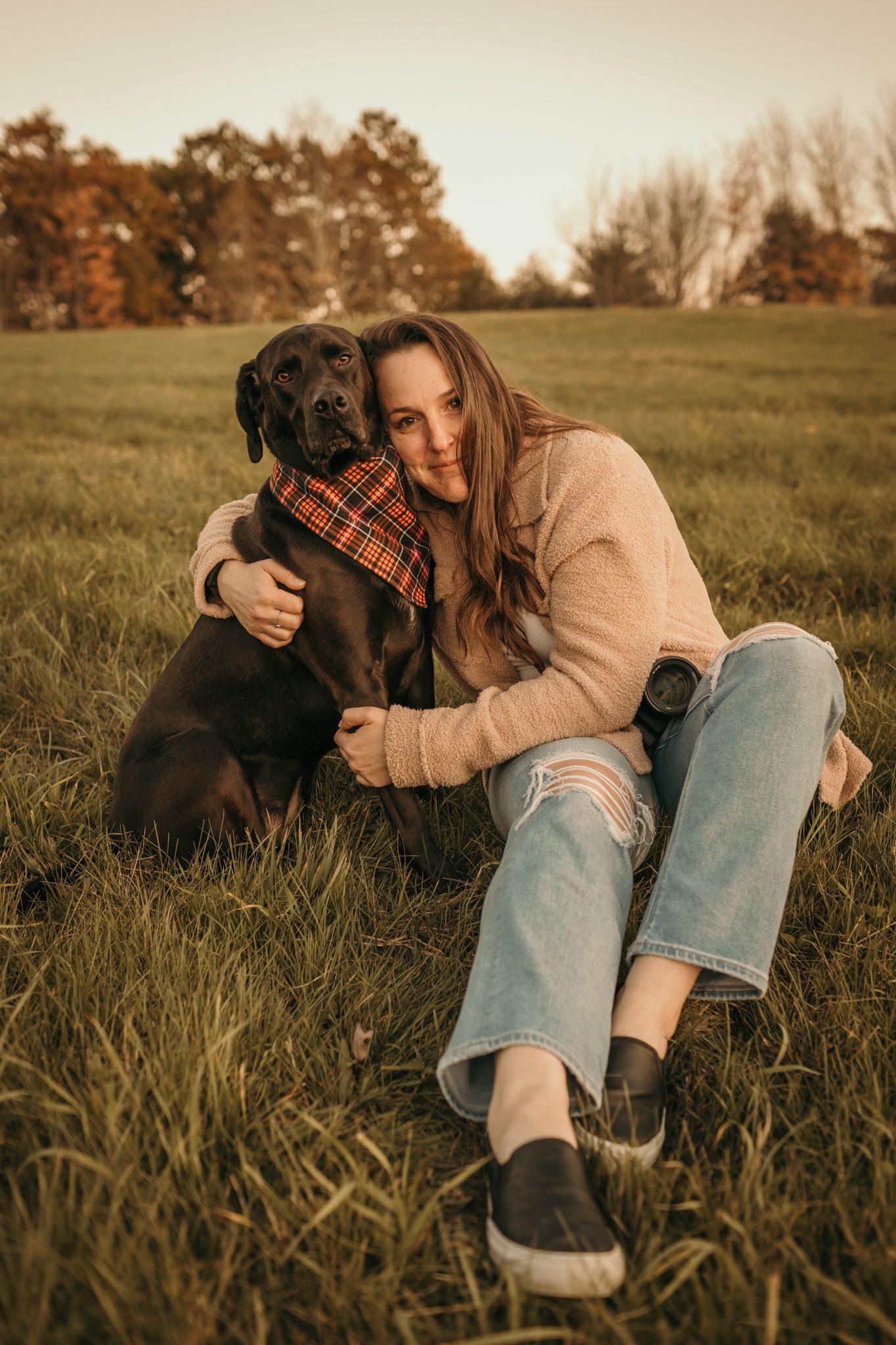 Person in casual attire hugging a black Labrador dog in an autumn field at sunset.