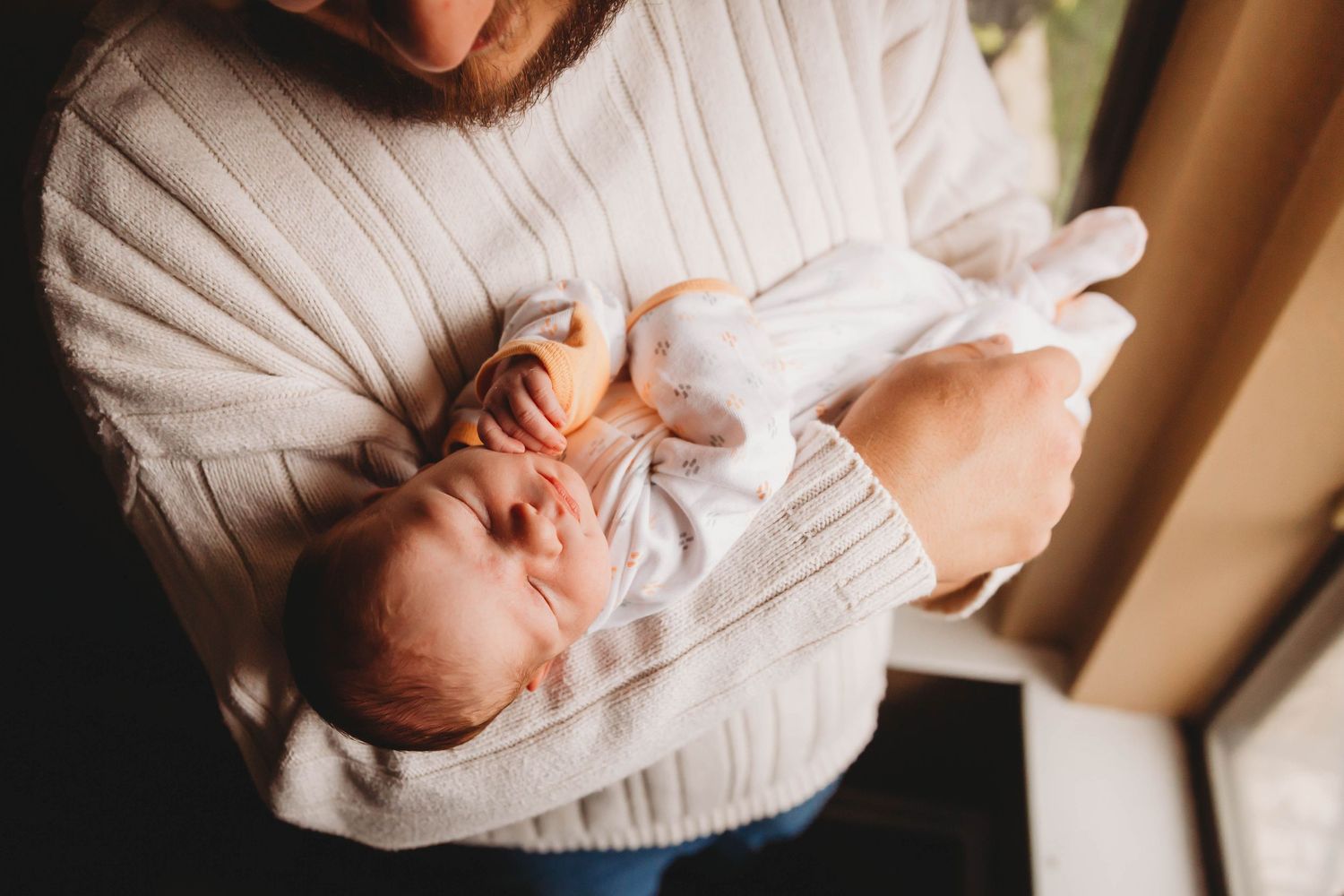 A parent in a white sweater cradles a tiny newborn baby swaddled in white.