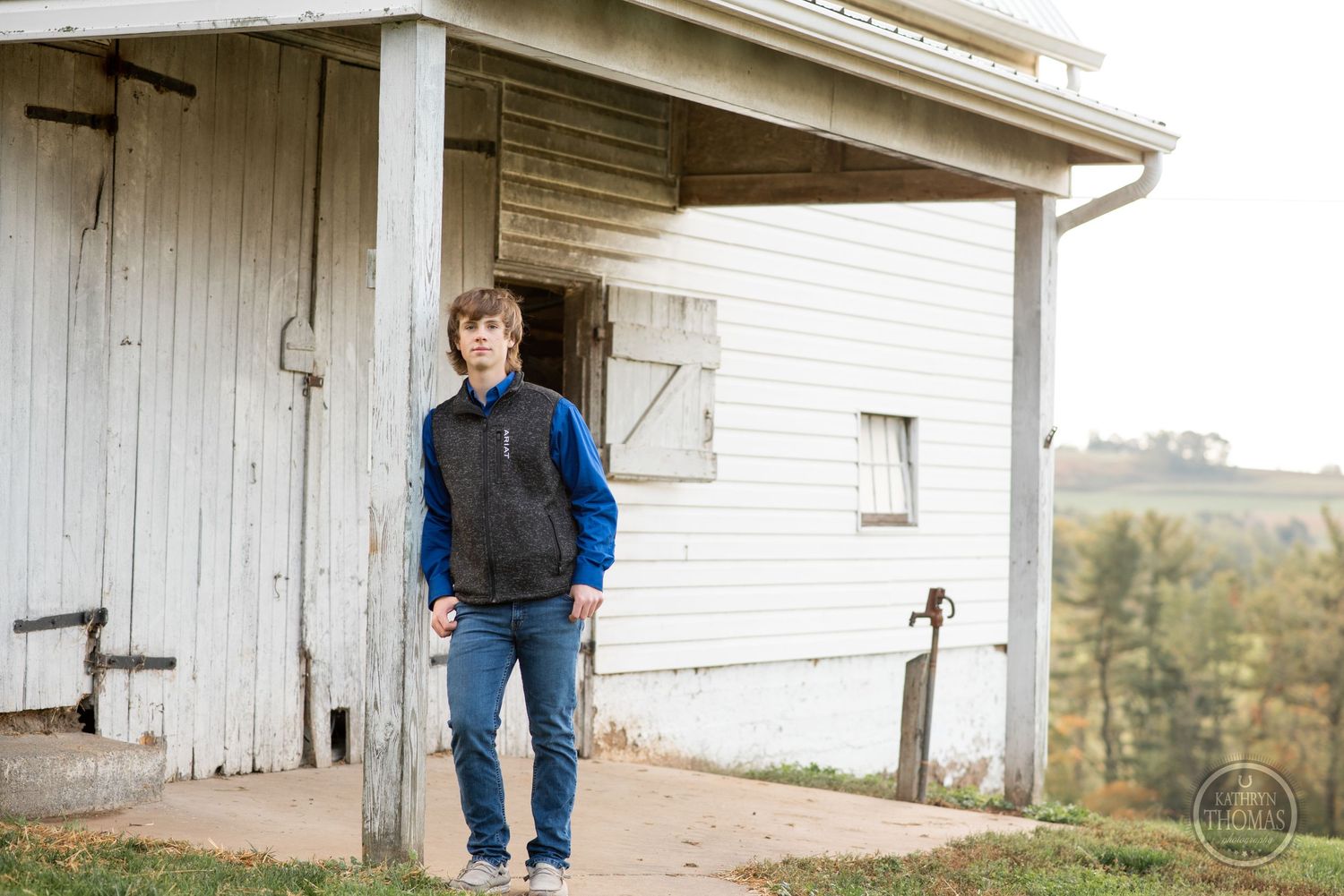 Figure in vest and jeans poses outside white barn with rural landscape in background.