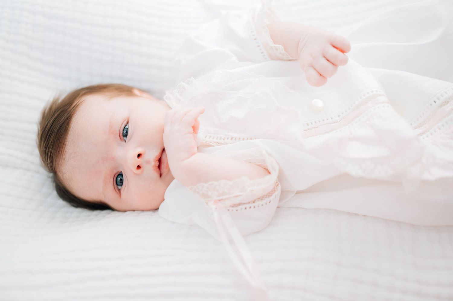 Sweet baby wearing white lying on white bedding looking up at camera.