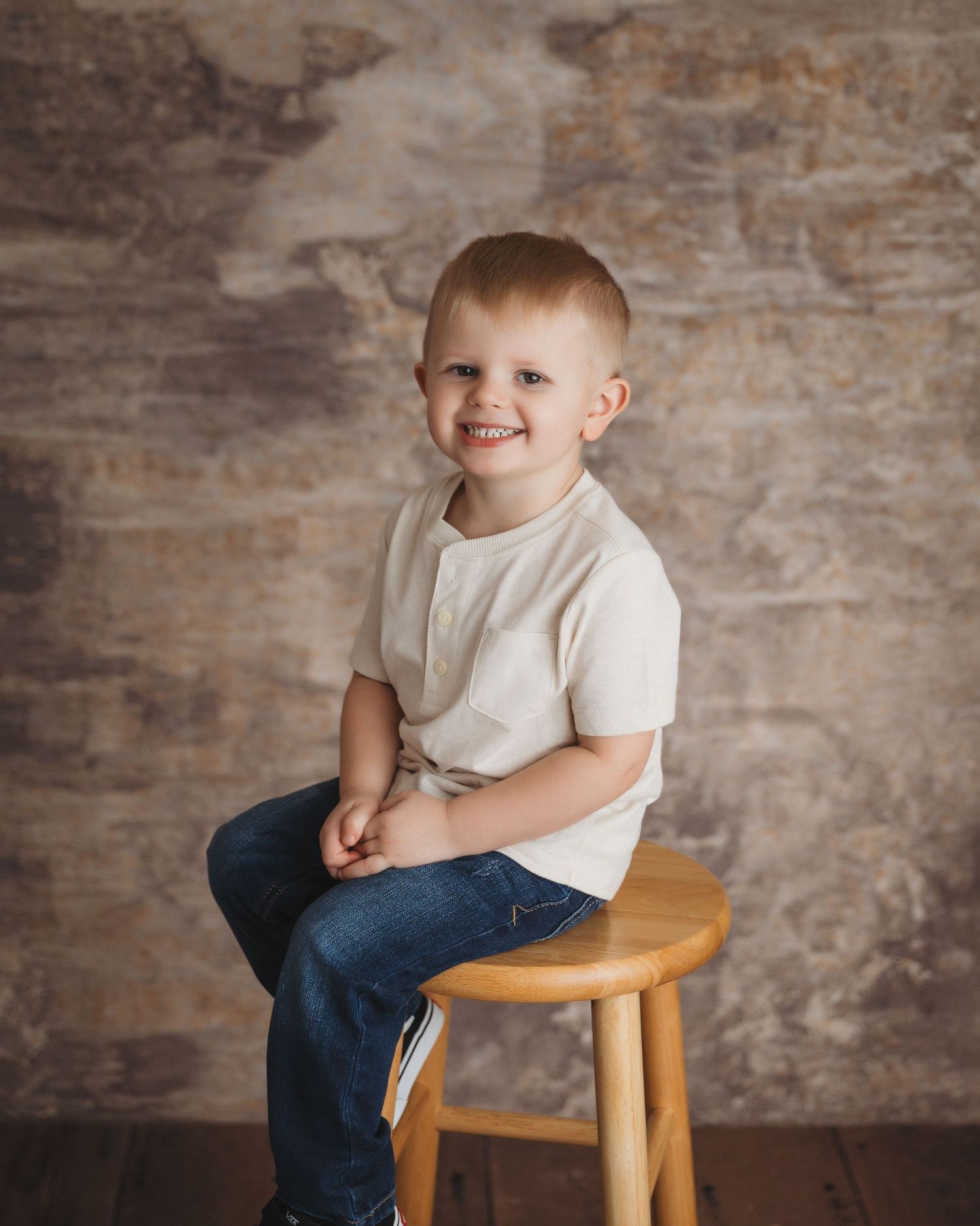 Young child in white t-shirt and jeans sitting on wooden stool against textured wall.
