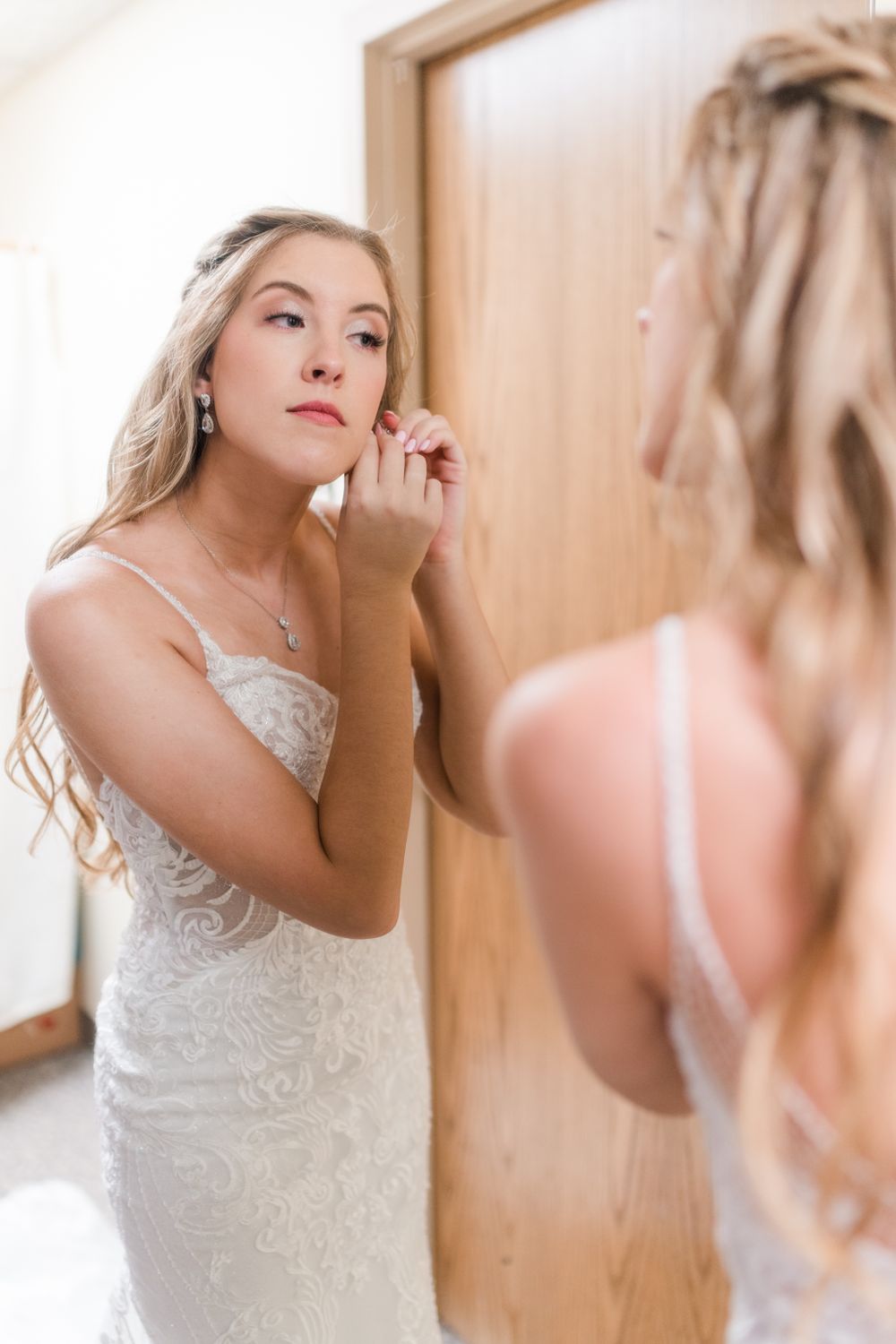 A reflection of someone putting on earrings while wearing a white lace wedding dress.