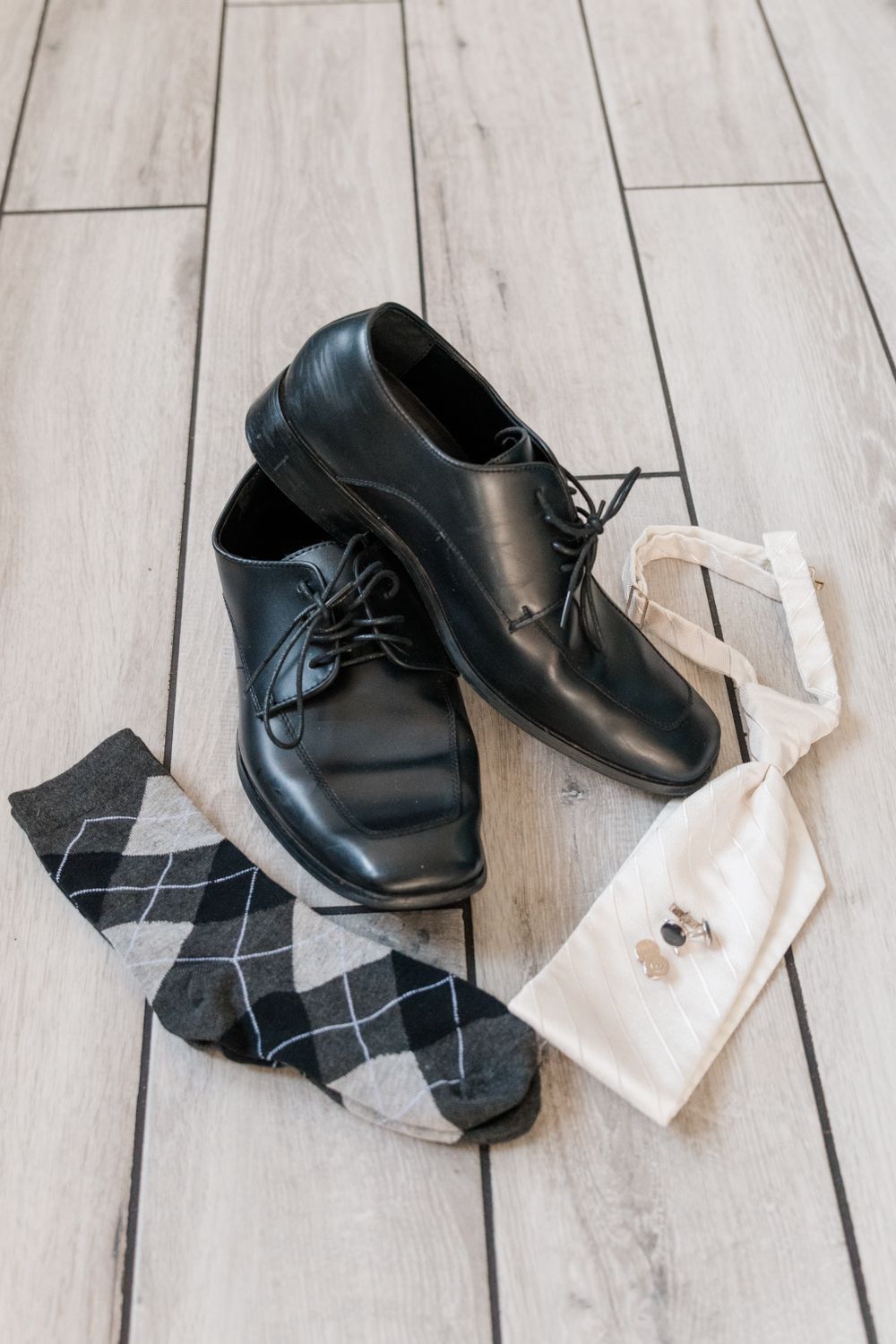 Black dress shoes with argyle socks and a white pocket square on wooden flooring.