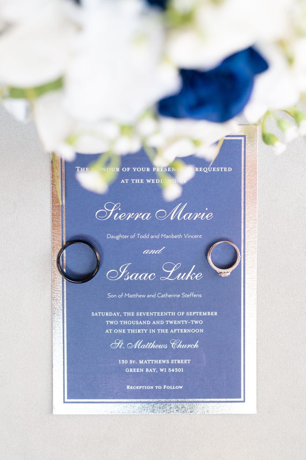 Navy blue wedding invitation with rings displayed on white floral background.
