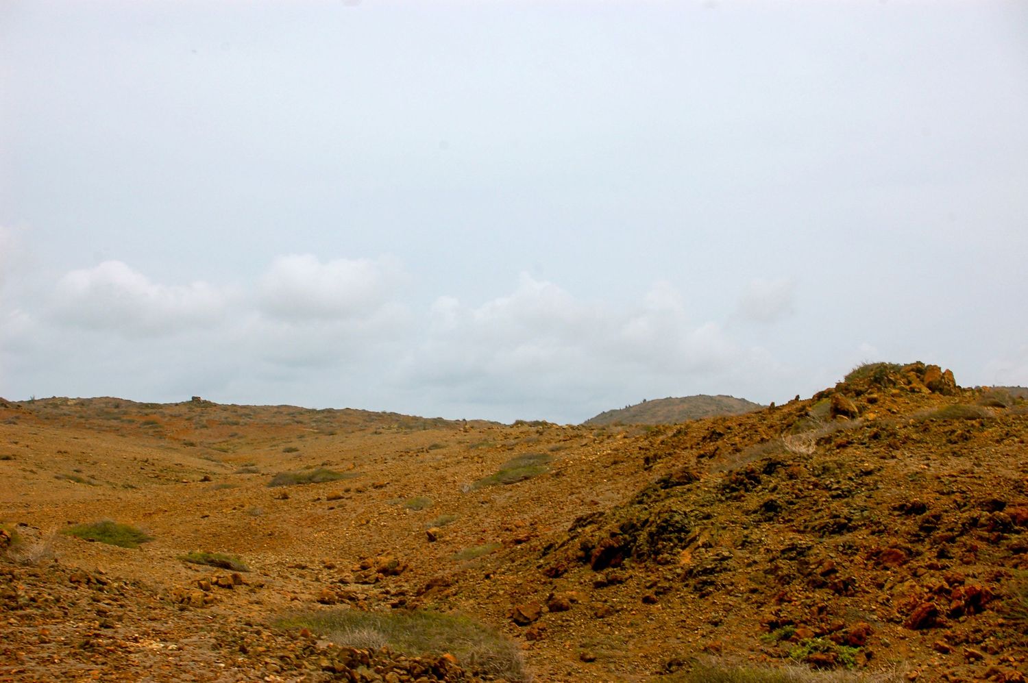Rolling desert hills under cloudy skies with reddish-brown soil and sparse vegetation.