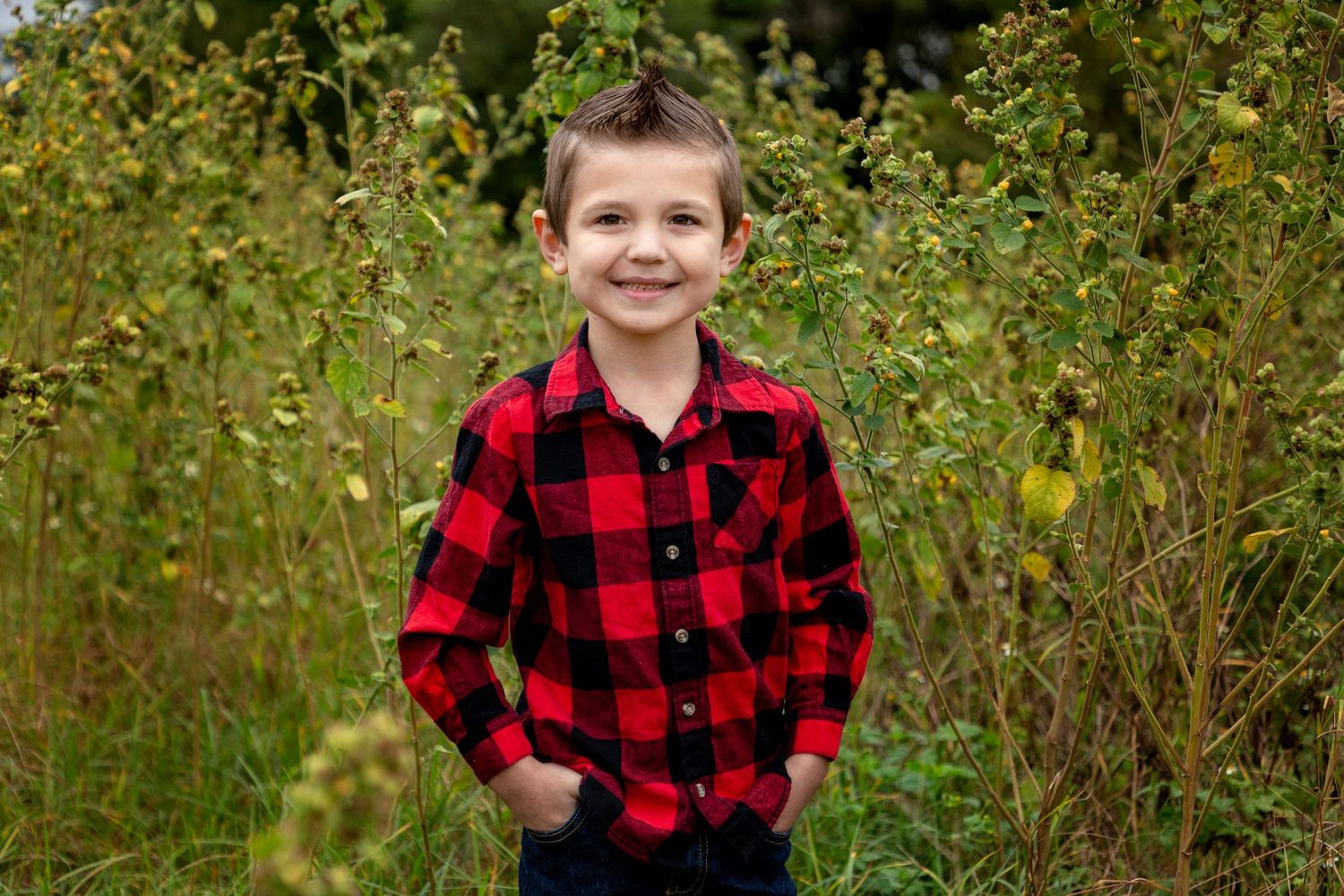 Little boy in a plaid shirt standing in the tall grass with his hands in his pockets.