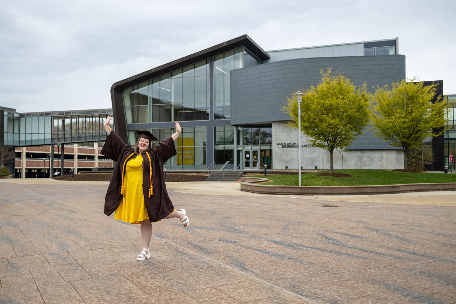 Modern glass and metal building exterior with person in graduation attire walking across plaza.