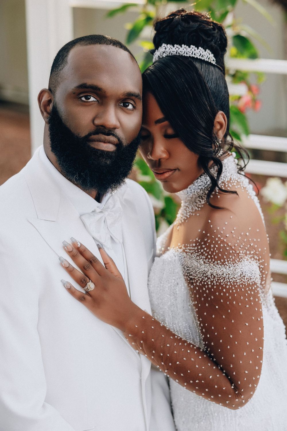 Groom in white suit and bride in pearl-embellished gown share intimate outdoor portrait pose.