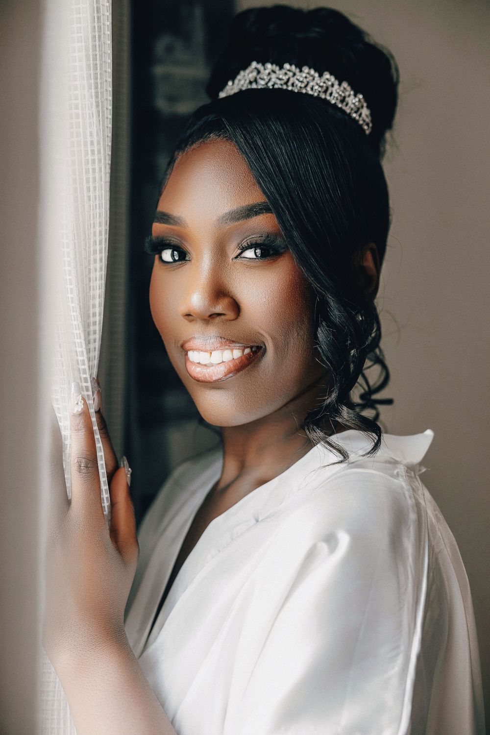Beautiful Black bride in white robe wearing crystal tiara smiles softly by window light.
