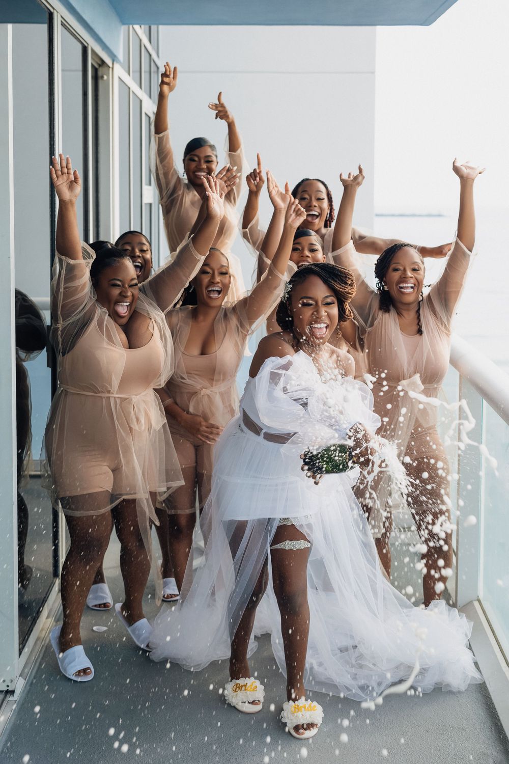 Excited bride and bridesmaids in nude dresses celebrate with confetti on hotel balcony.