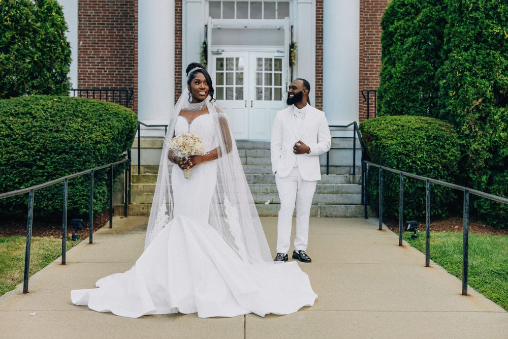 Black bride and groom in white wedding attire pose on church steps with manicured hedges.