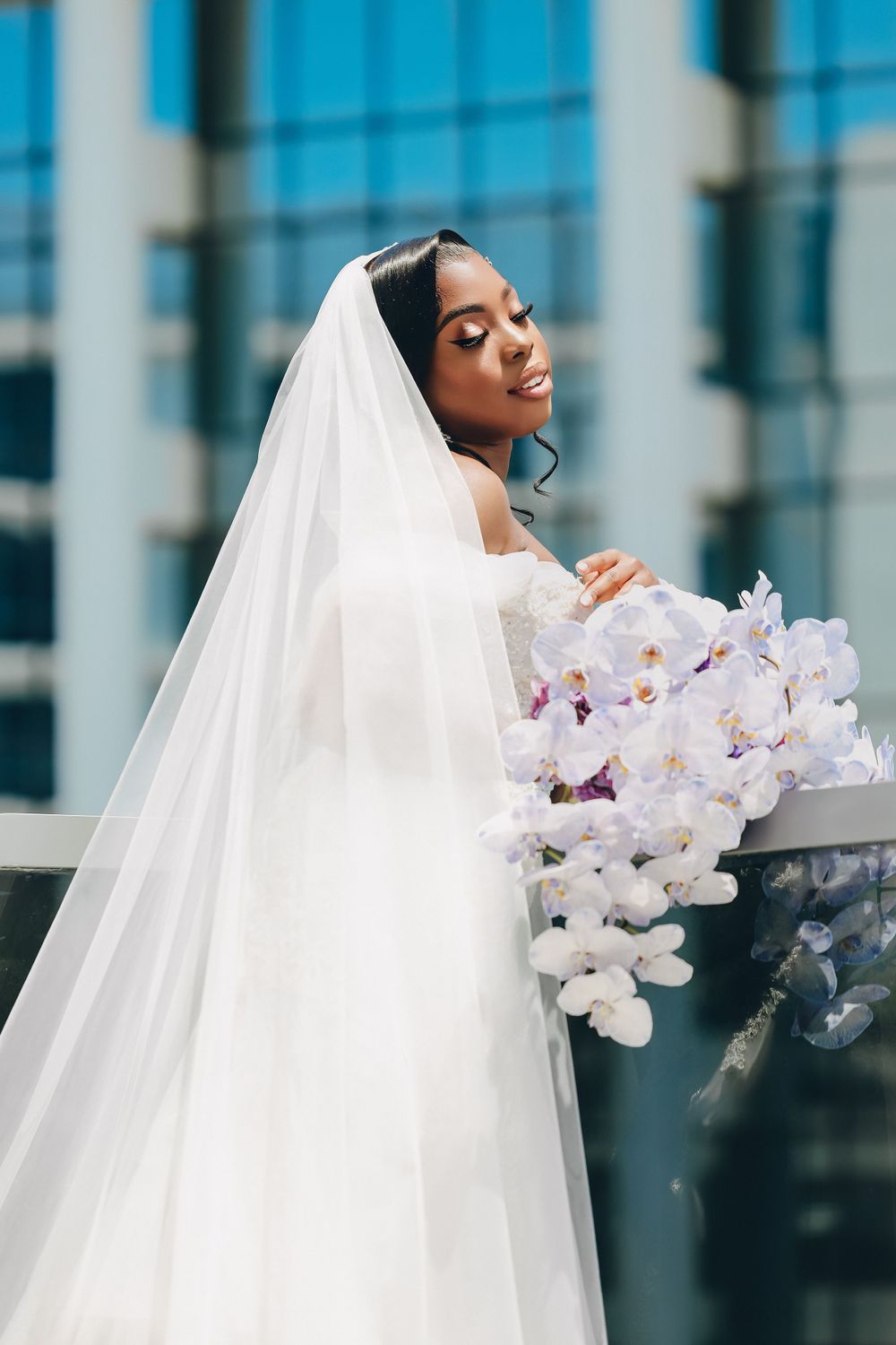 Beautiful Black bride in a white off-shoulder gown and veil holding white orchids on a balcony.
