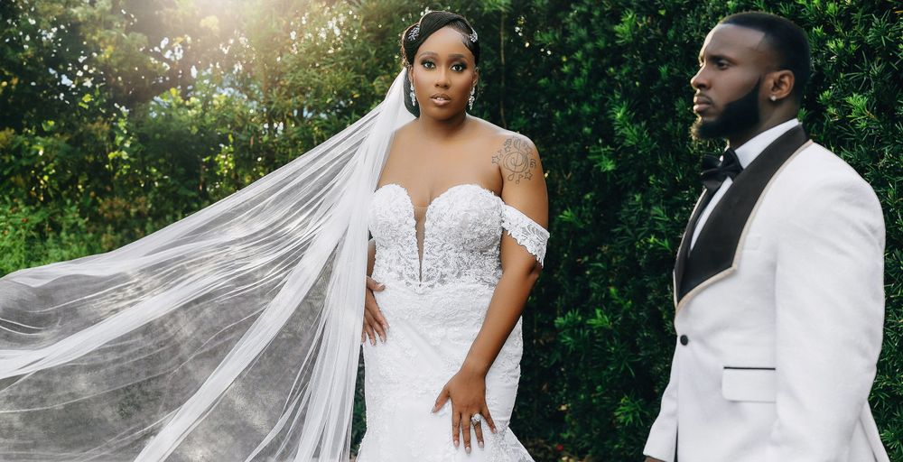 Black bride in beaded mermaid gown with long veil poses with groom in white tuxedo outdoors.
