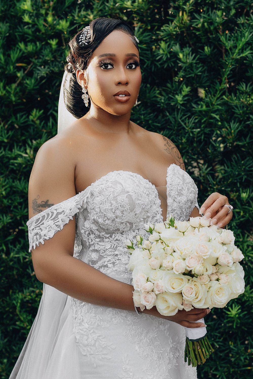 Bride in off-shoulder lace gown holds white rose bouquet before green hedge backdrop.