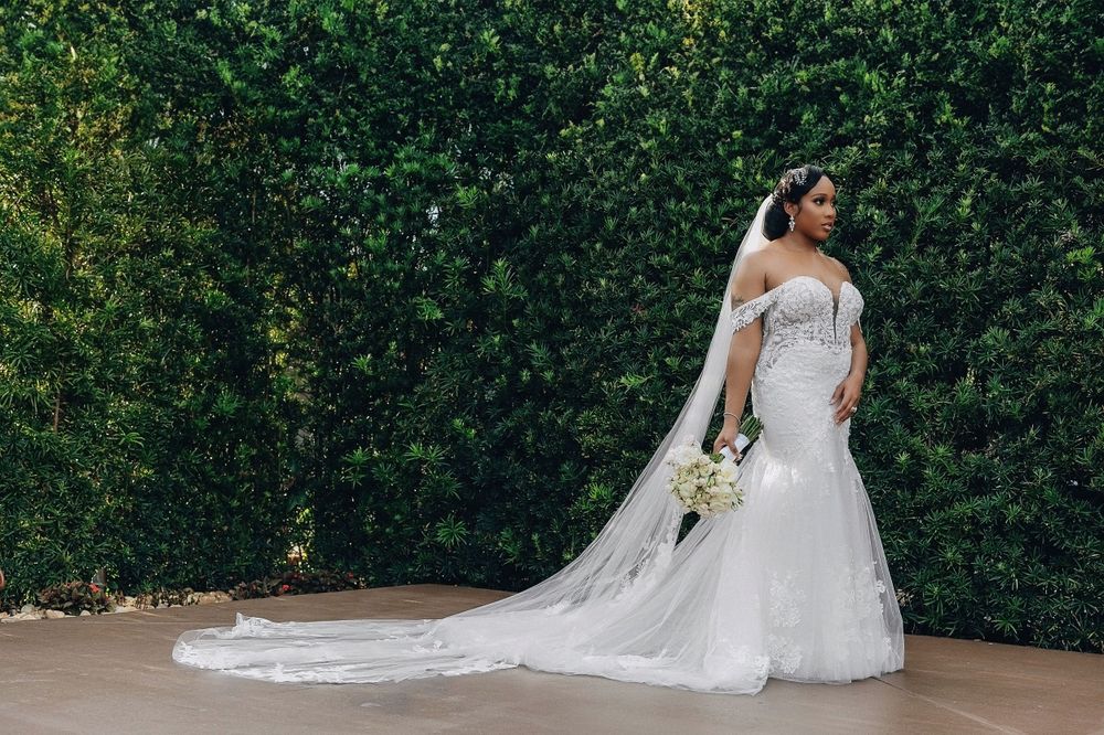Bride in off-shoulder mermaid gown with cathedral veil poses before lush green hedge wall.