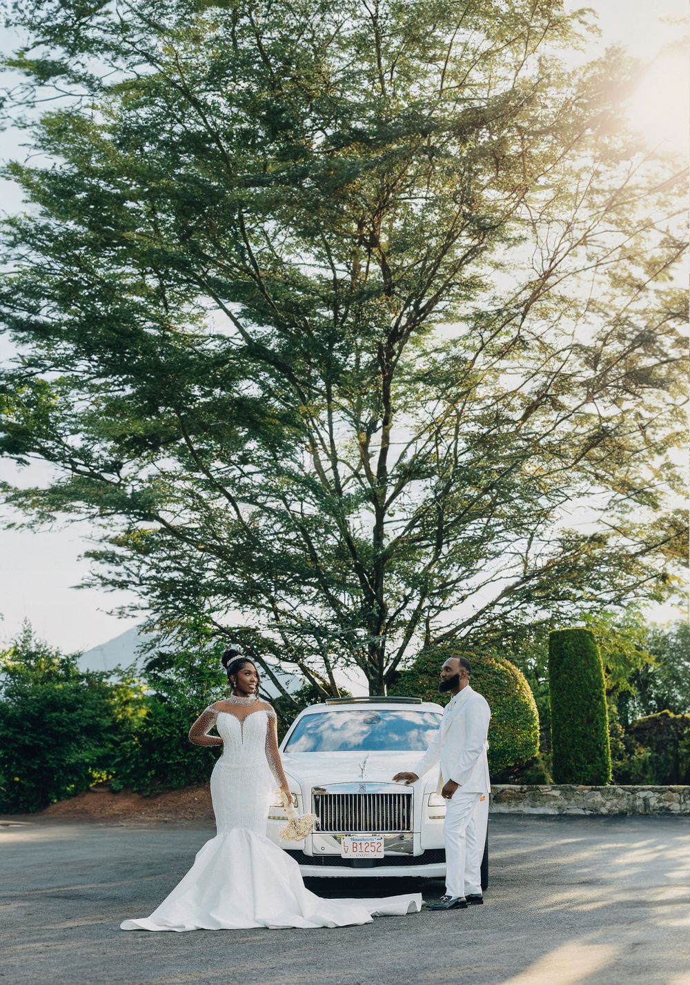 Black bride in a mermaid gown and groom in white standing before a white Rolls-Royce outdoors.