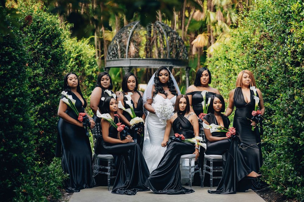 Bride in white gown poses with bridesmaids in black dresses beside ornate garden crown sculpture.