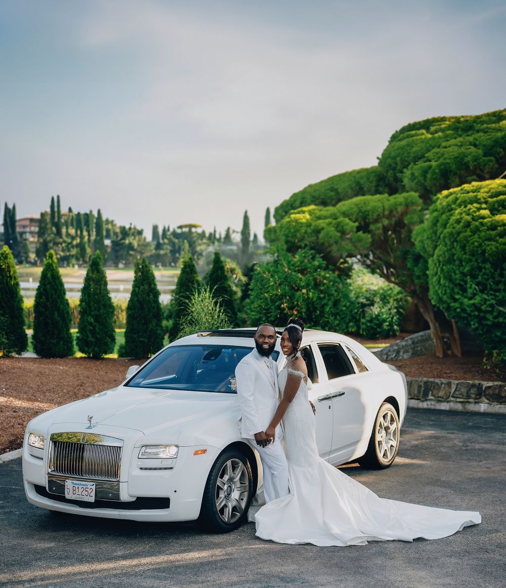Black bride and groom posing elegantly beside a white Rolls-Royce in a scenic outdoor setting.